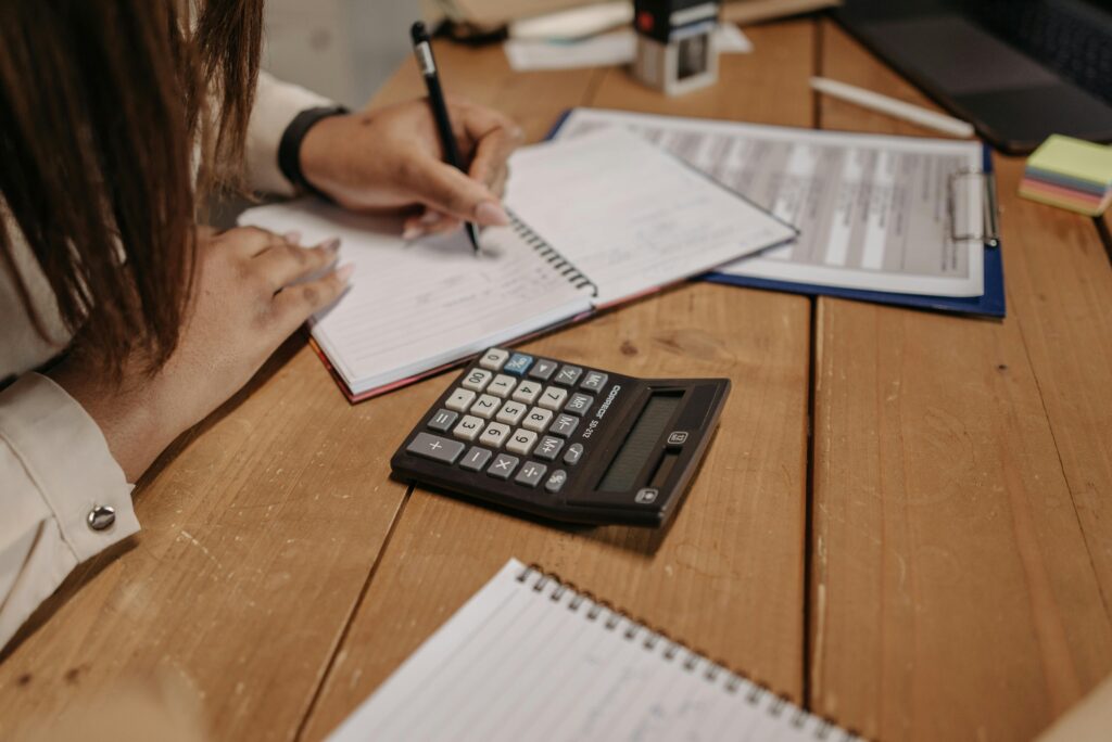 pexels-photo-7654576-7654576 Woman writing notes with a calculator on a wooden desk, focusing on work and accounting.