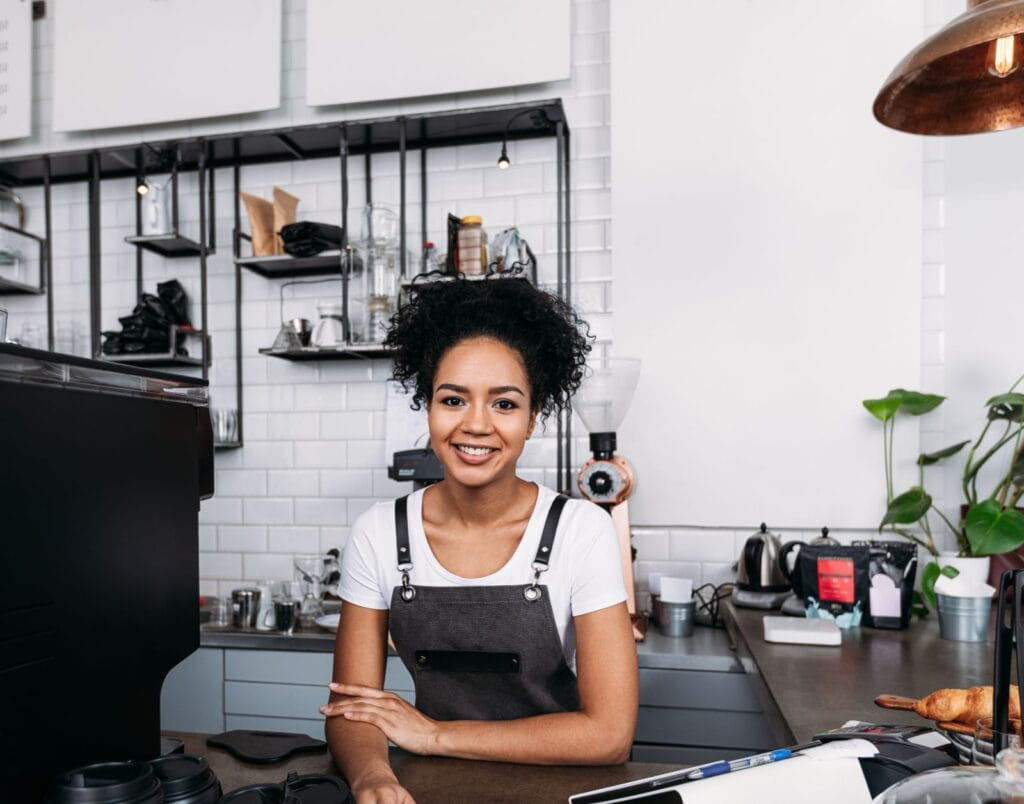 Barista wearing an apron stands behind a modern coffee shop counter, surrounded by equipment and plants, ready to serve customers.