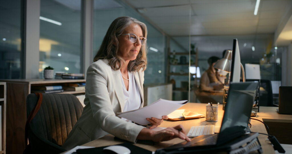 Businesswoman working late at a modern office desk, reviewing paperwork while using a computer and laptop, with colleagues visible in background.