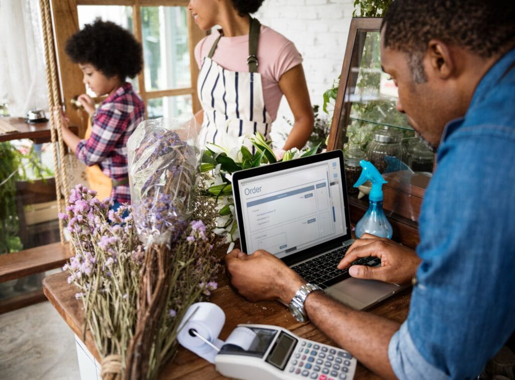 Florist processing an online order at the shop counter, using a laptop beside flowers, payment terminal, and coworker helping a customer at the door.