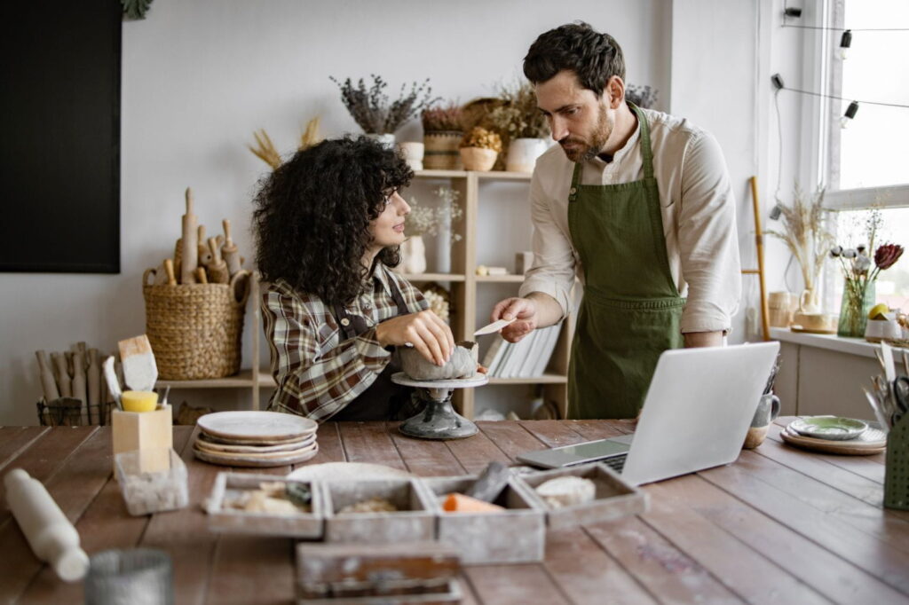 Instructor guiding a student in a pottery class, shaping clay together at a wooden table with tools, plates, and a laptop nearby.