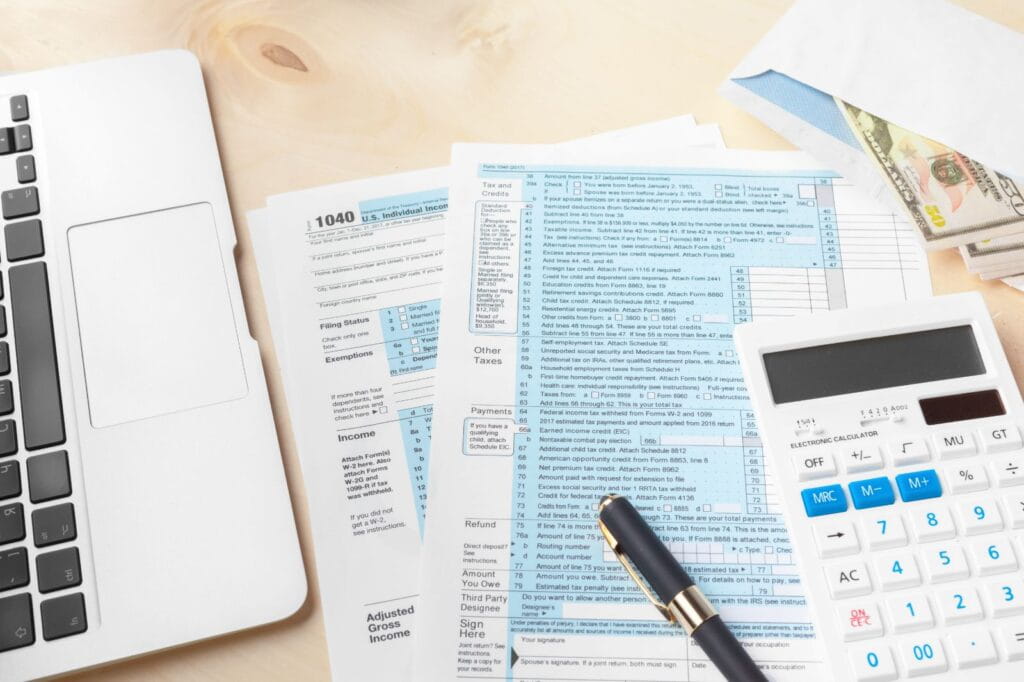 Laptop, tax forms, calculator, pen, and cash-filled envelope arranged on a desk, representing personal income tax preparation and financial planning.