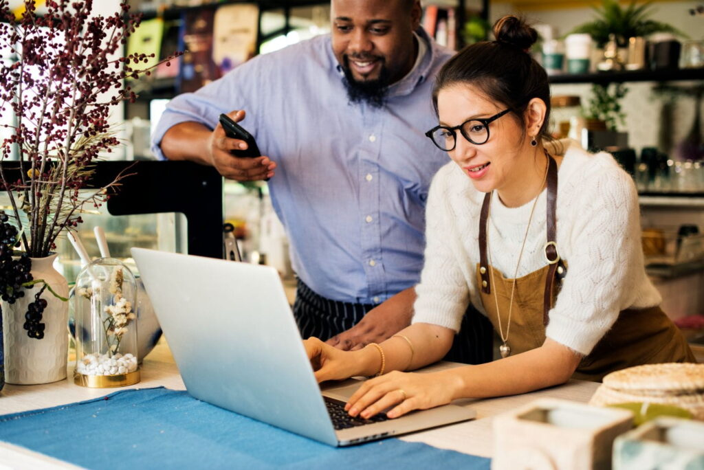 Small business owners collaborating at a counter, smiling while reviewing something on a laptop and smartphone inside a cozy, modern shop.