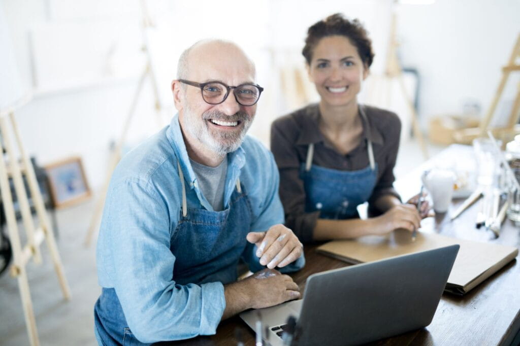 Two artists wearing aprons sit together at a studio table, collaborating on a laptop surrounded by creative tools and sketchbooks.