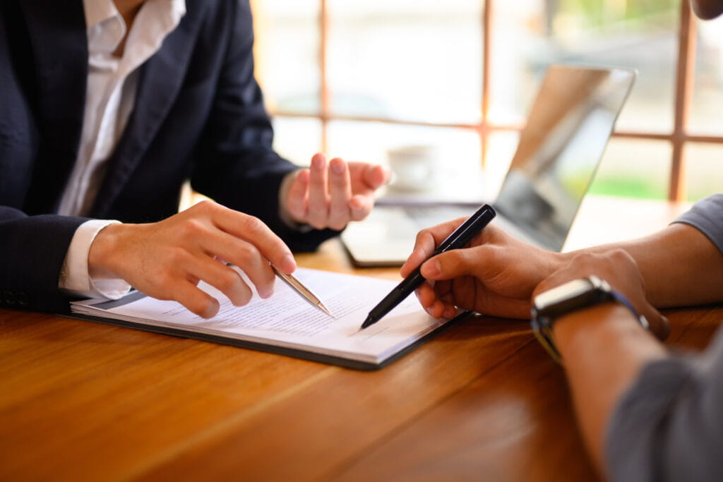 Two professionals sit at a wooden table, reviewing and signing a document together with pens, laptop and coffee in the background.