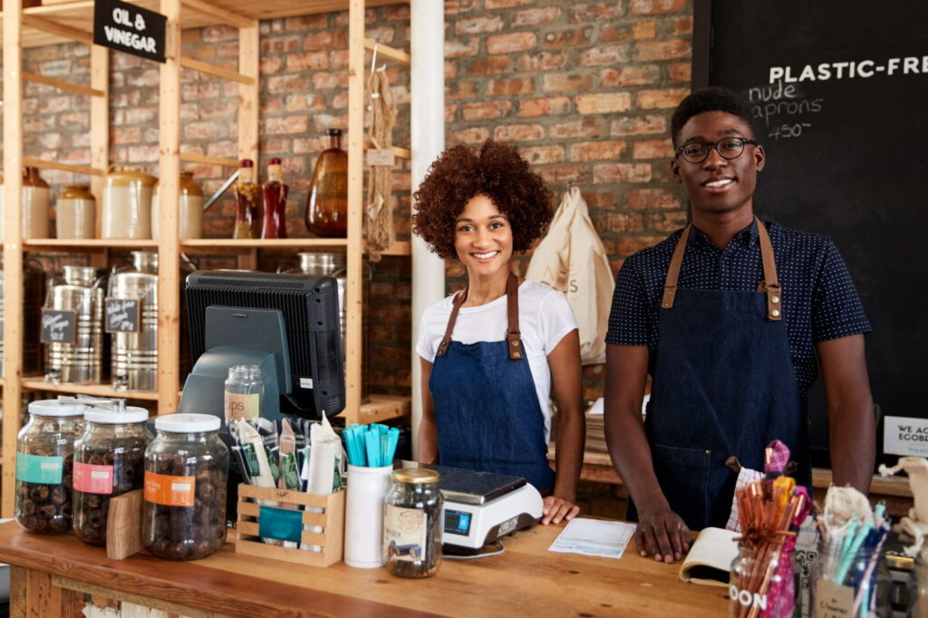 Two shop employees wearing denim aprons stand behind a wooden counter in a cozy zero-waste store with jars and reusable products. Two shop employees wearing denim aprons stand behind a wooden counter in a cozy zero-waste store with jars and reusable products.