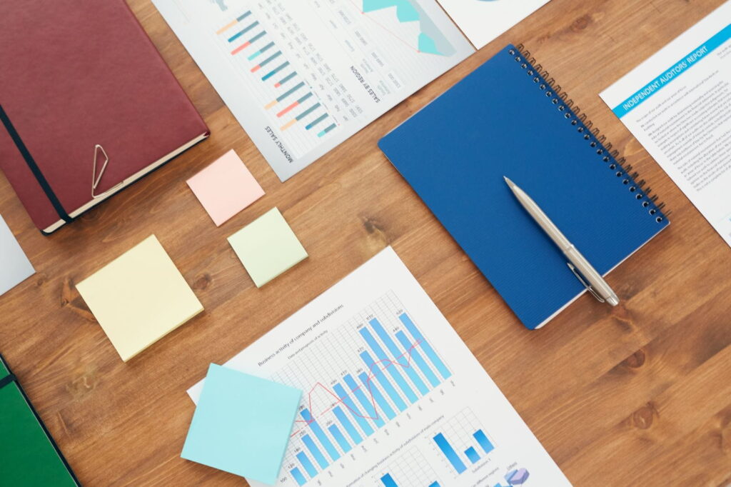 Wooden desk with financial charts, colorful sticky notes, notebooks, and a pen neatly arranged, representing business planning, analysis, and office organization.